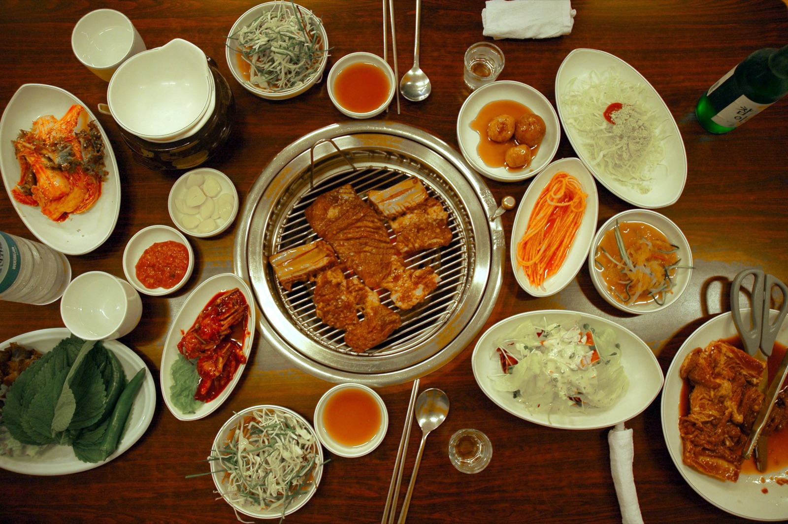 Overhead view of a Korean barbecue table with beef short ribs on a round charcoal grill surrounded by kimchi, salads, dips, perilla leaves, green chilli and a pair of scissors