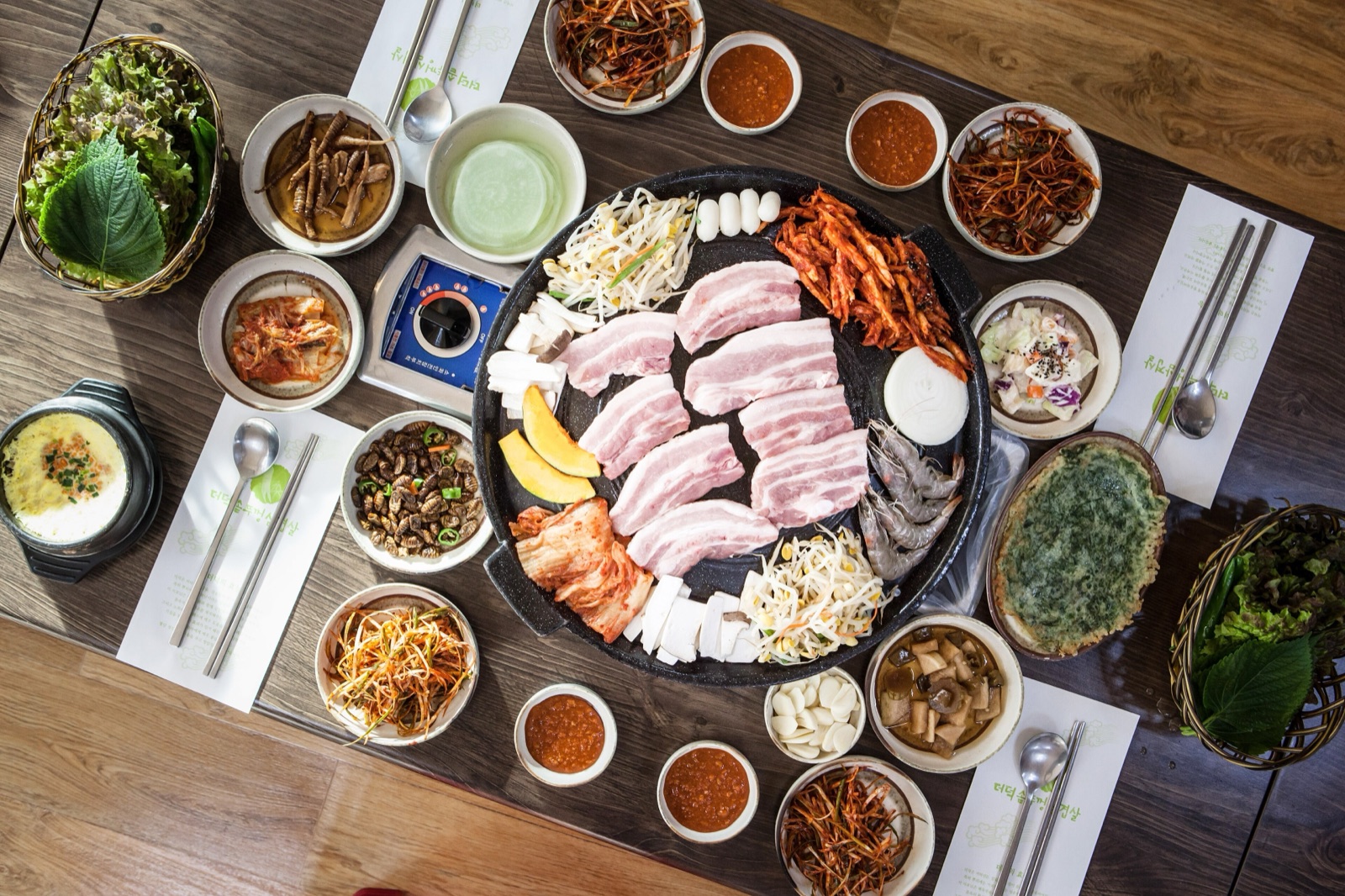 Overhead view of a Korean barbecue table with raw pork belly on a round grill plate surrounded by kimchi, bean sprouts, dips, lettuce and perilla leaves