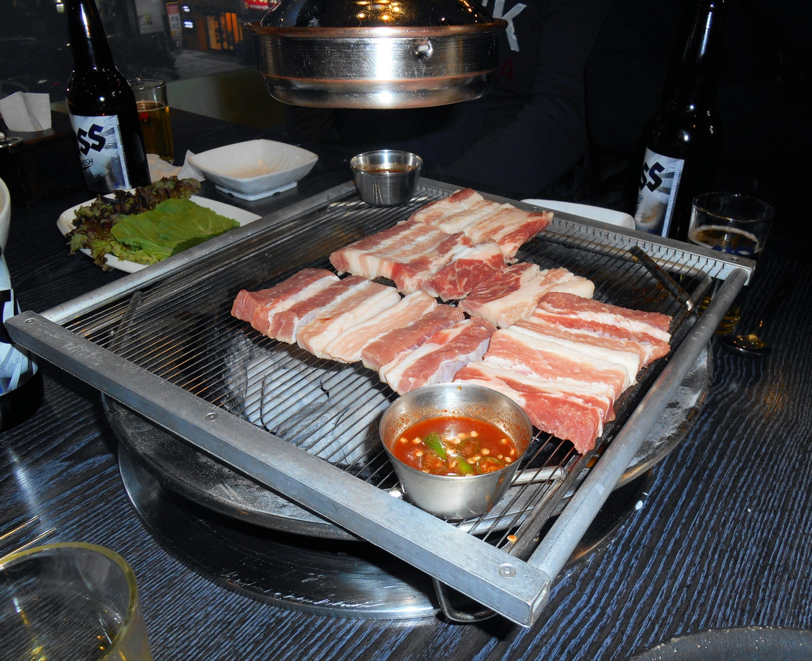 Raw pork on a charcoal grill set into a restaurant table in Changwon, South Korea, with a metal extractor hood above, a plate of lettuce and bottles of beer