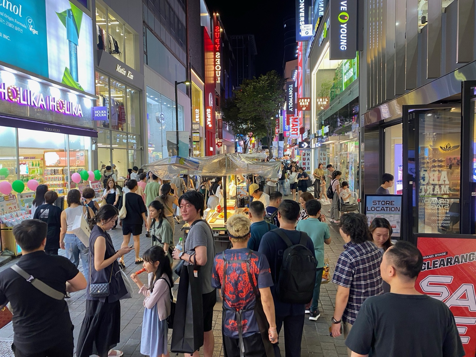 夜のソウル・明洞。明るい看板の下の通りを多くの買い物客が歩き、屋台が並んでいる