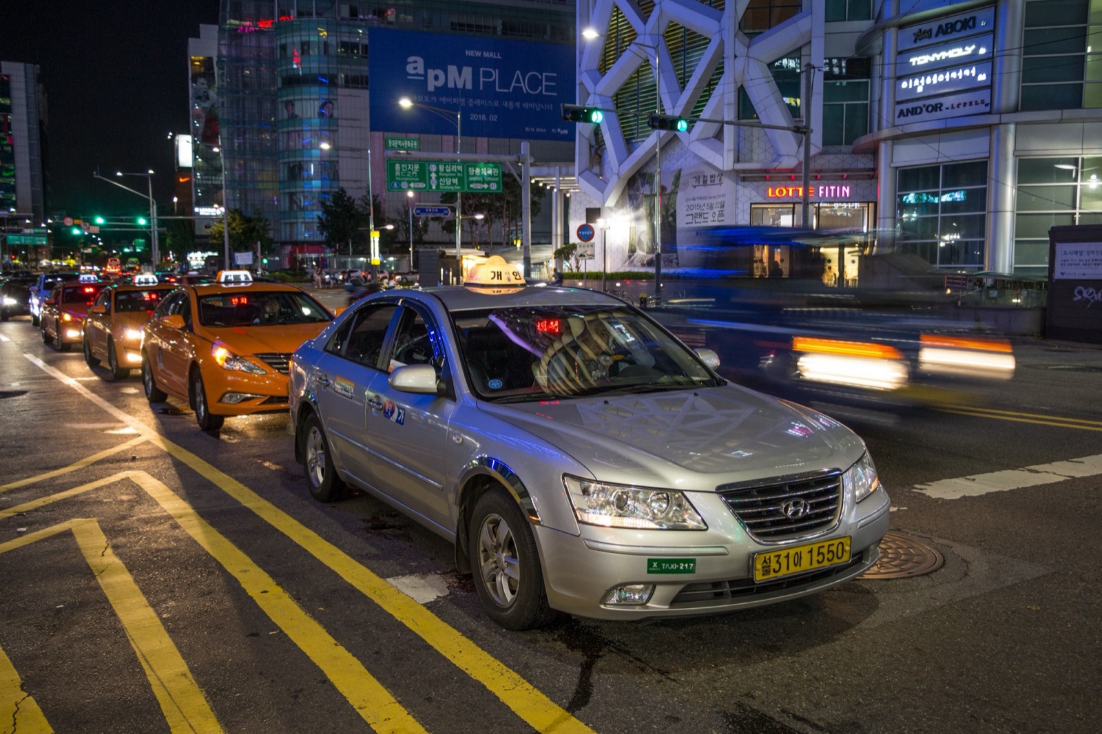 夜のソウルで、明るく照らされたショッピングモールの前の道路に、銀色のタクシーとオレンジ色のタクシーが並んでいる