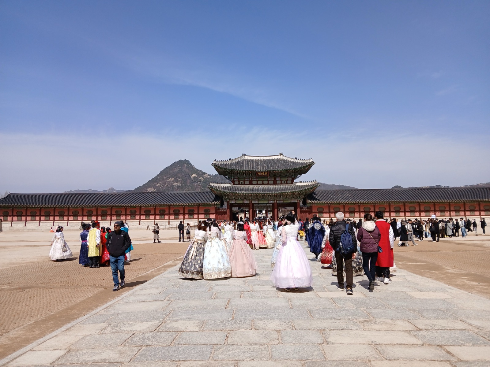 Visitors in colourful rented hanbok walk across the stone courtyard toward Heungnyemun Gate at Gyeongbokgung Palace in Seoul, with Bugaksan mountain behind