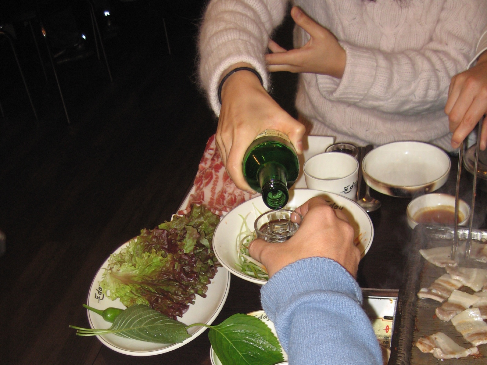 韓国式焼肉の席で、女性が向かいの人の小さなグラスに焼酎を注いでいる