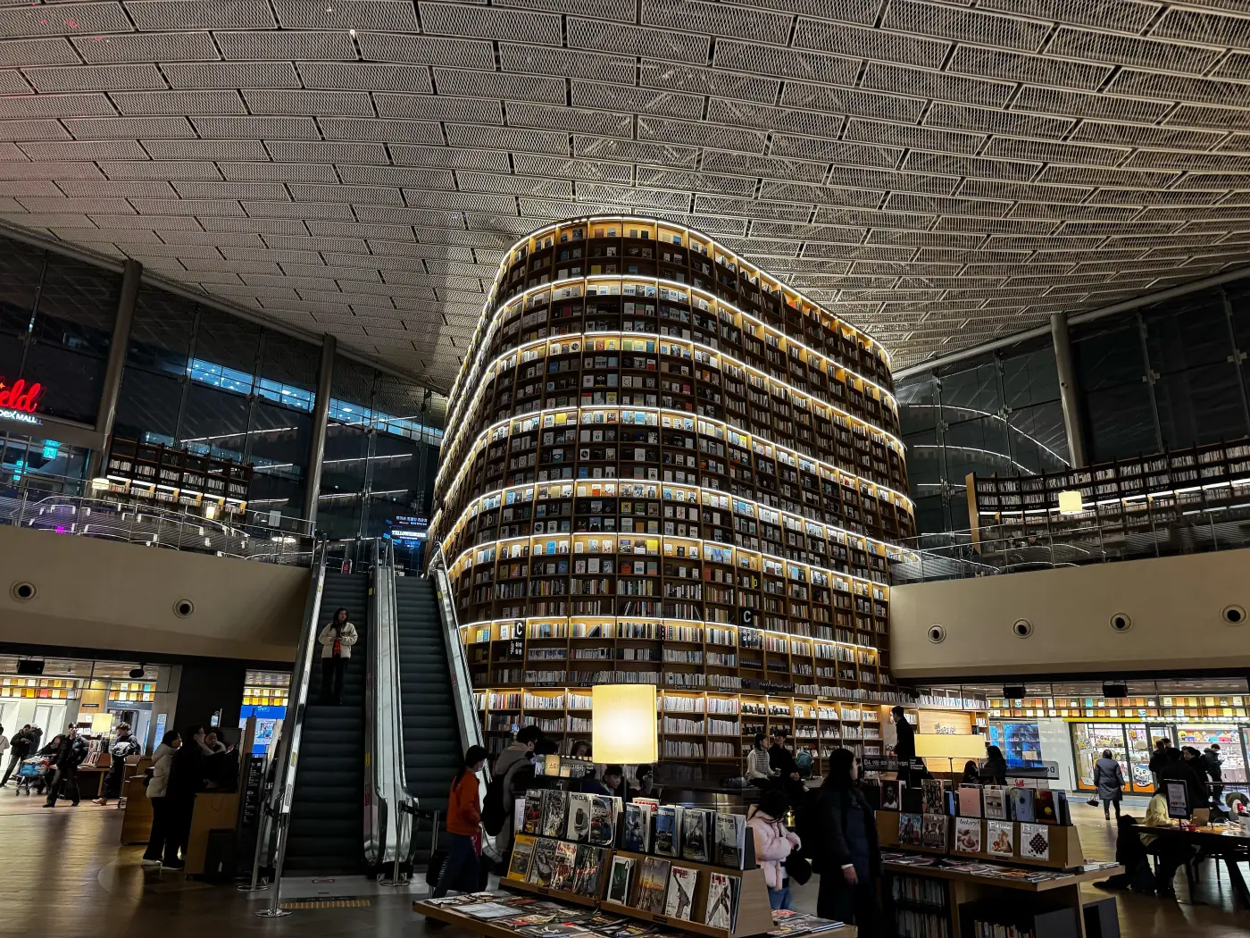 Tall curved bookshelves lit from within rise above escalators and shoppers at Starfield Library inside COEX Mall, Seoul