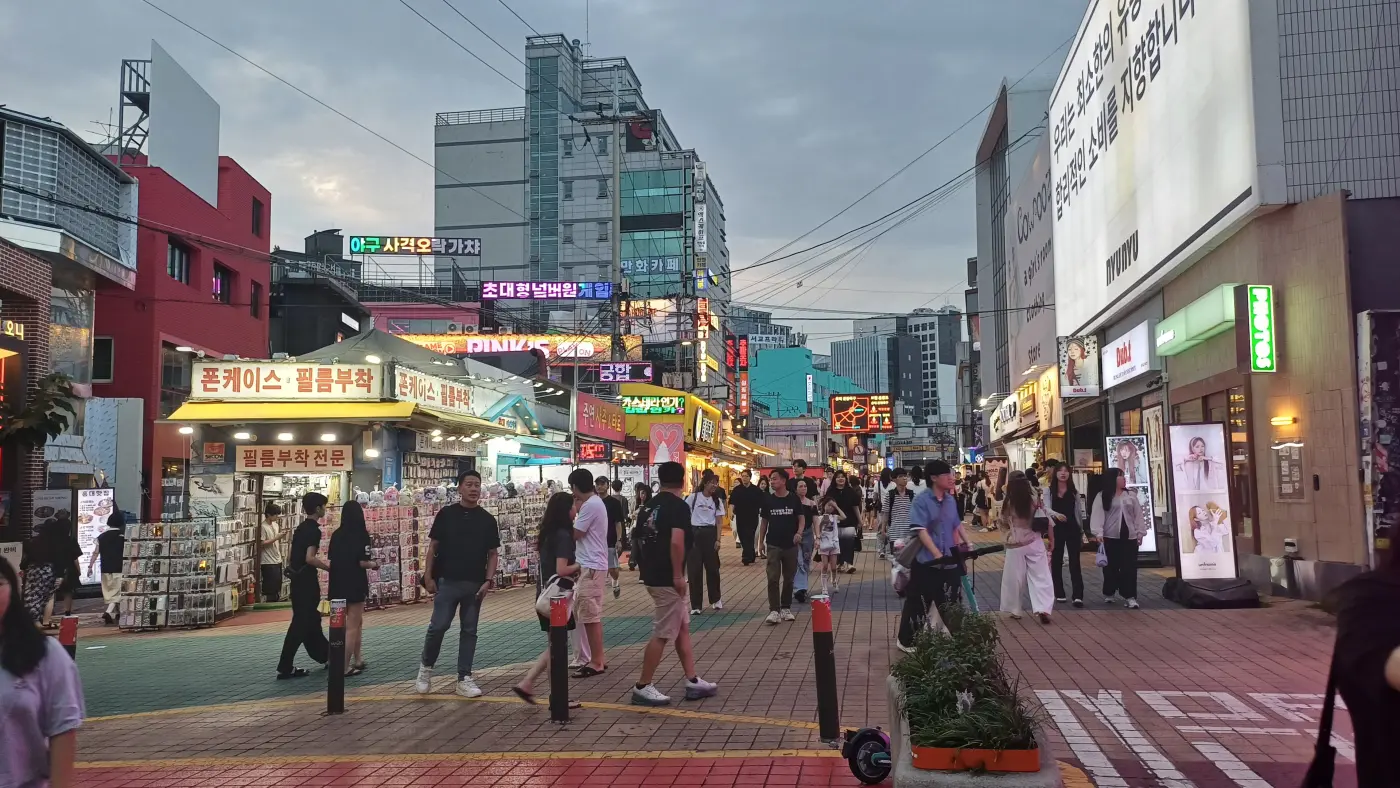 A pedestrian street in Hongdae, Seoul, at dusk, lined with phone-case shops, neon signs and people strolling
