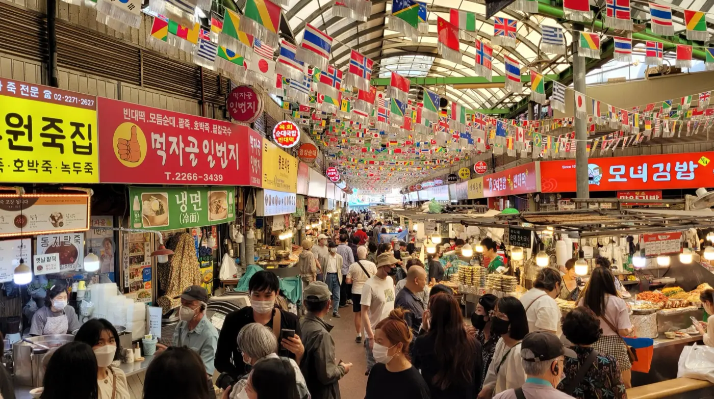 ソウル・広蔵市場の中、万国旗の下に色とりどりの看板を掲げた食べ物の店が並び、大勢の人が行き交う