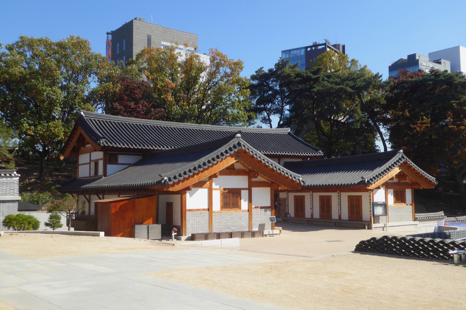 白い壁と瓦屋根の平屋の伝統家屋が、晴れた日の中庭に建っている。背後には紅葉した木々と現代的なオフィスビルが見える